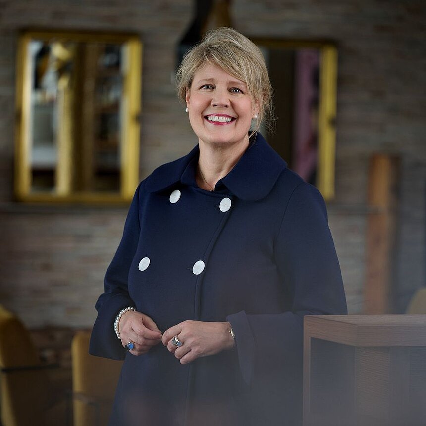 Lächelnde Frau mit blonden Locken in dunkelblauer Jacke steht in einem modernen Raum mit Spiegeln im Hintergrund