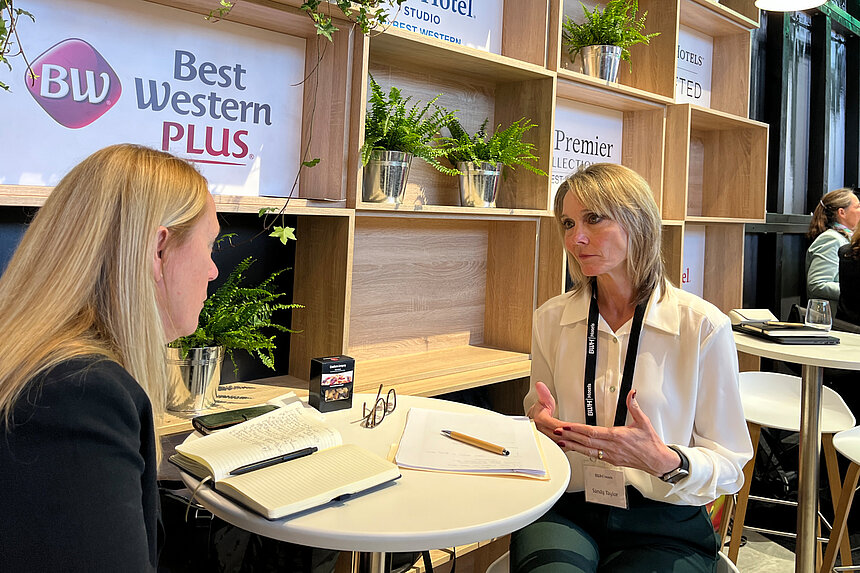Zwei Frauen im Gespräch an einem runden Tisch auf einem Messestand von Best Western Plus, mit Notizbuch und Pflanzen im Hintergrund.