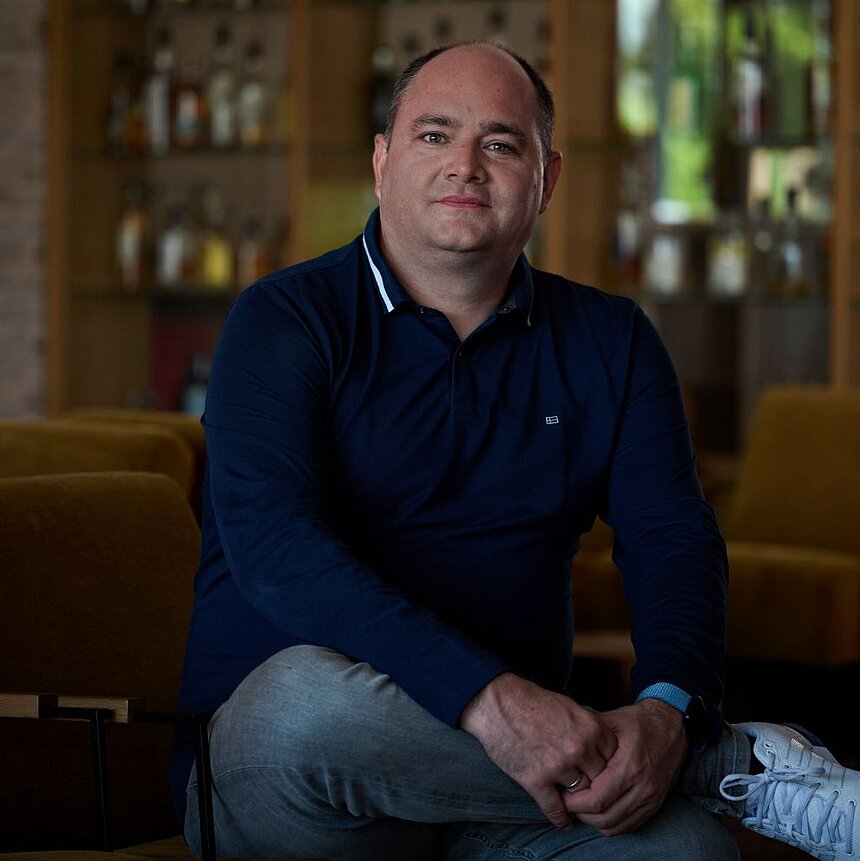 ChatGPT: Mann mit kurzgeschnittenem Haar in dunkelblauem Langarmshirt sitzt mit verschränkten Händen in einem modernen Raum mit Regalen im Hintergrund