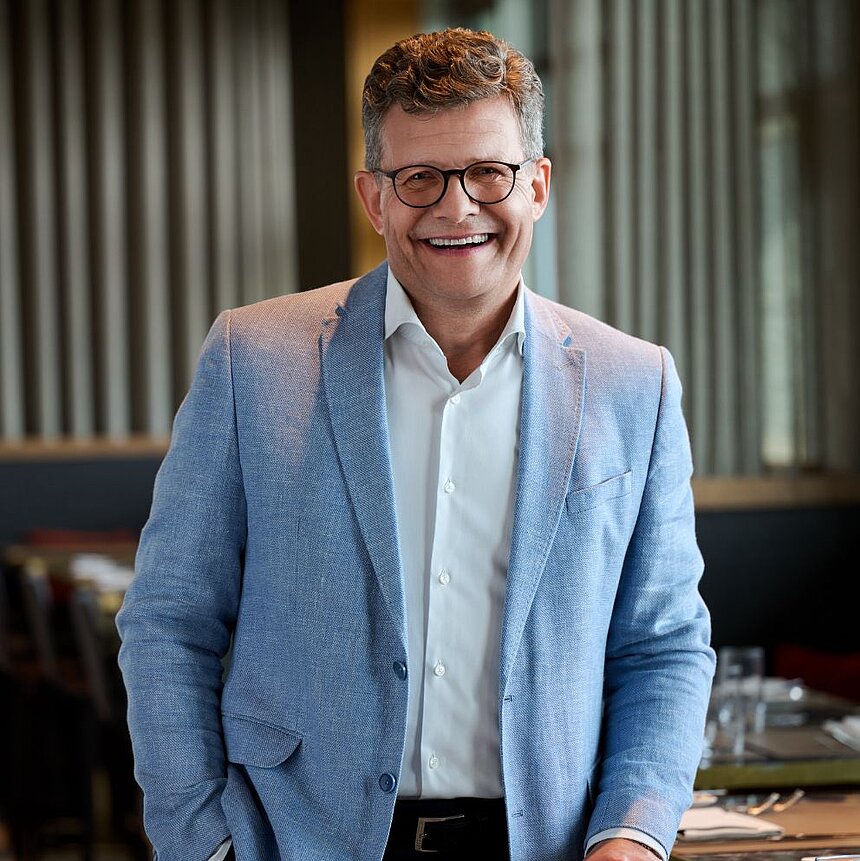 ChatGPT: Lächelnder Mann mit Brille in hellblauem Sakko und weißem Hemd steht in einem modern eingerichteten Raum