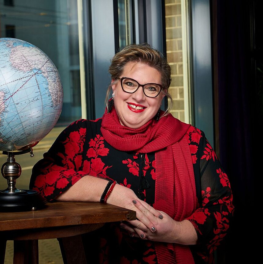 Lächelnde Frau mit Brille in schwarzem Kleid mit rotem Muster und rotem Schal sitzt neben einem Globus an einem Holztisch