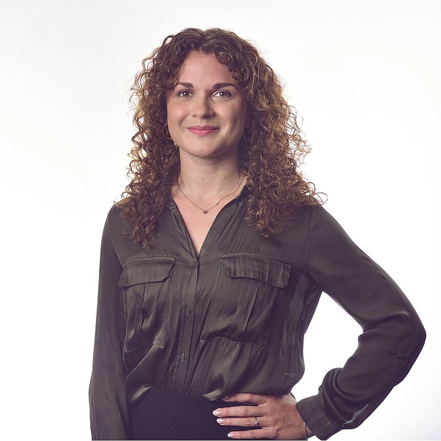 Frau mit lockigen Haaren in dunkelgrüner Bluse steht mit einer Hand in der Hüfte vor hellem Hintergrund