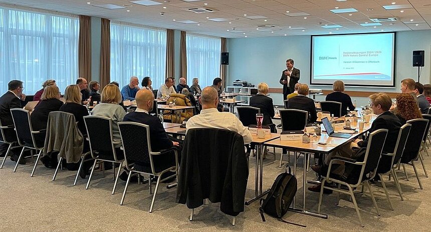 Teilnehmer einer Konferenz sitzen in einem Tagungsraum an Tischen, während ein Redner vorne vor einer Präsentationsfolie spricht